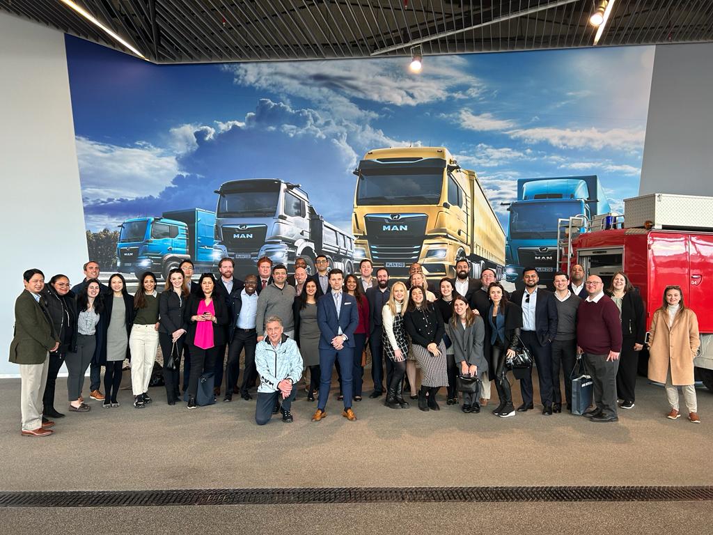 A diverse group of professionals posing for a group photo in front of a wall mural featuring three MAN trucks in blue, yellow, and teal colors at the MAN Truck and Bus SE headquarters in Germany. The group consists of men and women of various ages and ethnicities, dressed in business and casual attire.