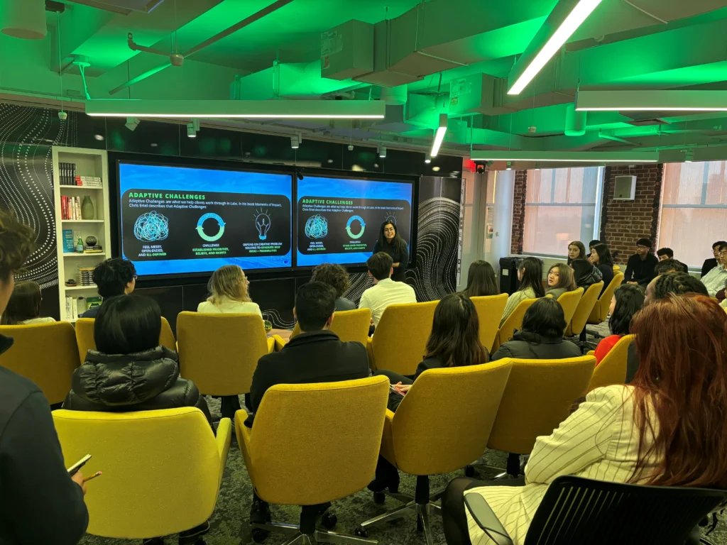 Deloitte AI Wharton conference in NYC.Presentation on adaptive challenges with an audience in a modern conference room. Presenter stands in front of two large screens displaying slides about adaptive challenges, surrounded by attendees seated in yellow chairs.