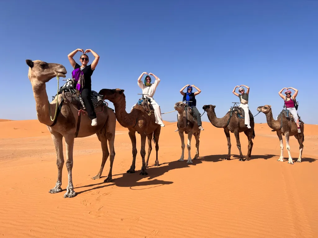 Group of women riding camels in the Sahara Desert, making heart shapes with their hands, under a clear blue sky on the orange sand dunes.