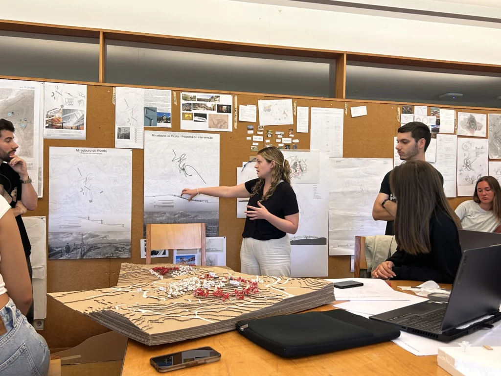 Belmont University presenting in Portugal. A group of architecture students engaged in a design critique session, discussing project proposals with maps, drawings, and a 3D model displayed on a table.