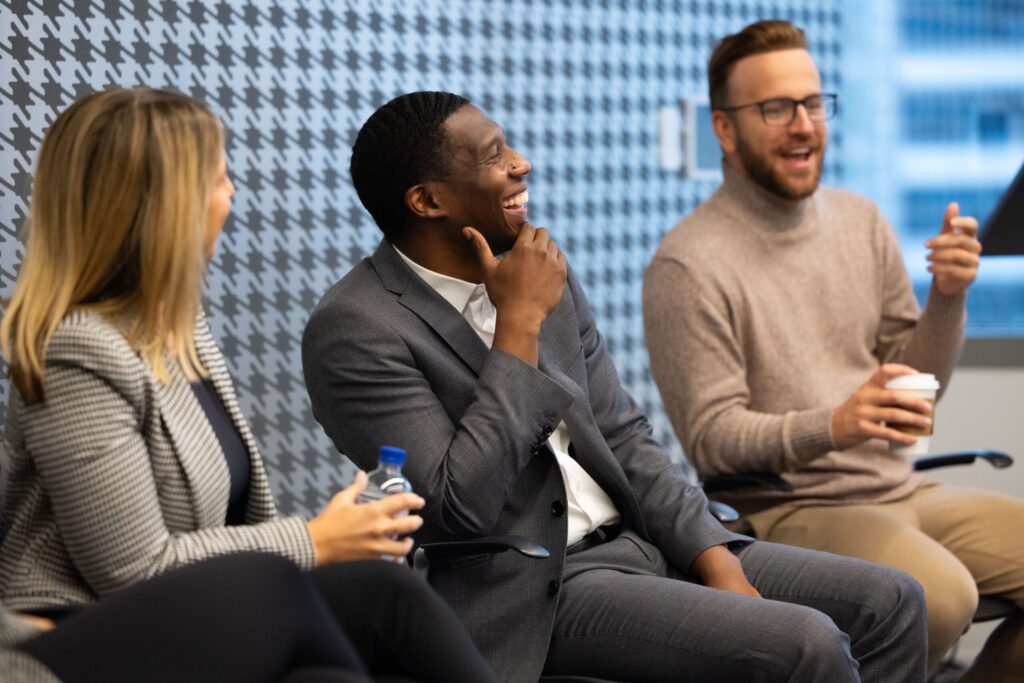 Three business professionals engaging in a lively discussion during a panel session in an office setting. One man in a grey suit is laughing, while another man in glasses and a beige sweater is speaking, holding a coffee cup, and a woman in a checkered blazer is smiling and listening attentively.