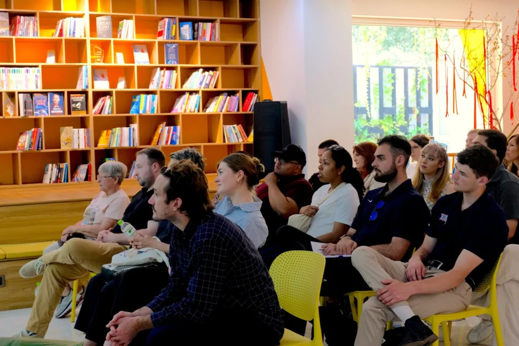 A diverse audience attentively listening to a presentation in a modern library setting, with colorful bookshelves in the background, creating an atmosphere of learning and engagement.