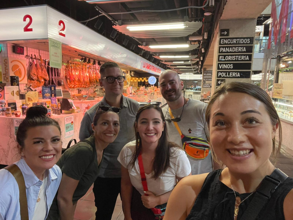 A group of six smiling friends or colleagues taking a selfie inside a vibrant market in Madrid, Spain, with various signs and a display of hanging cured meats in the background, indicating a lively atmosphere filled with local delicacies. University of Oregon in Madrid market.