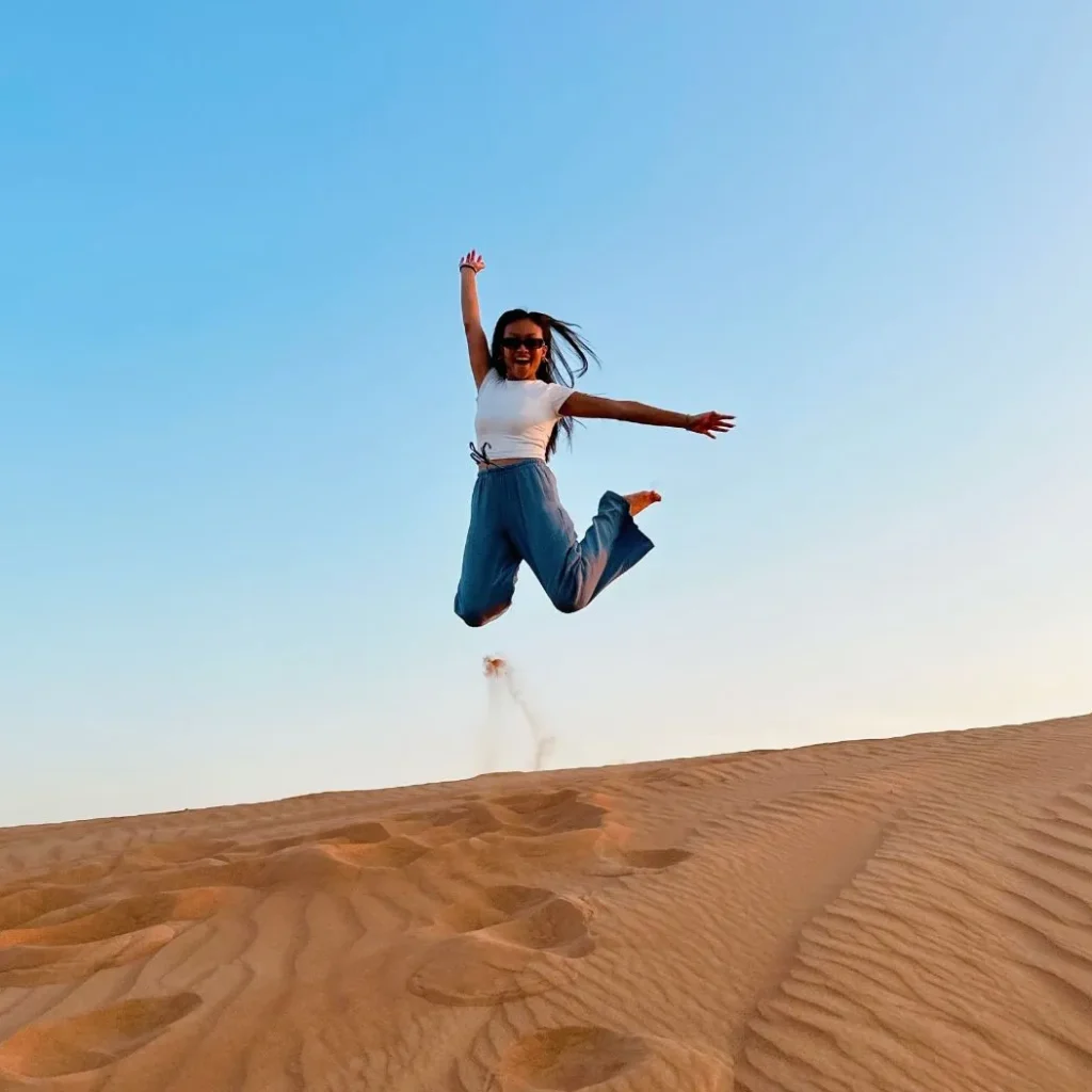 A joyful woman jumping mid-air on a sand dune in the desert at sunset, showcasing freedom, adventure, and happiness against a clear blue sky.