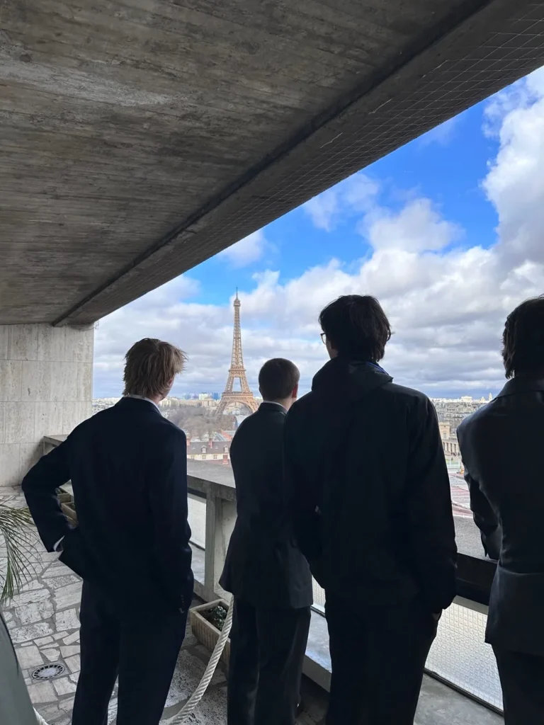 Three young men in suits standing on a balcony, looking at the Eiffel Tower on a cloudy day in Paris, symbolizing ambition, travel, and inspiration.