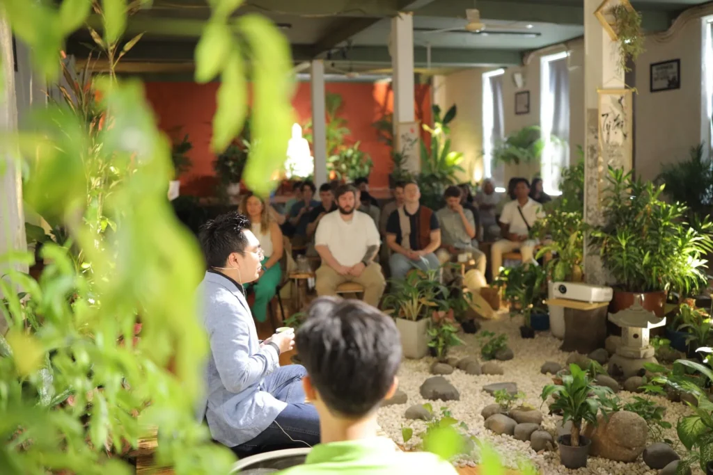 A group of people seated in a tea house in Vietnam, surrounded by lush green plants and natural decor, attentively listening to a speaker in a light blue blazer. The setting has a calming ambiance with potted plants, pebbles, and natural light streaming in through the windows.