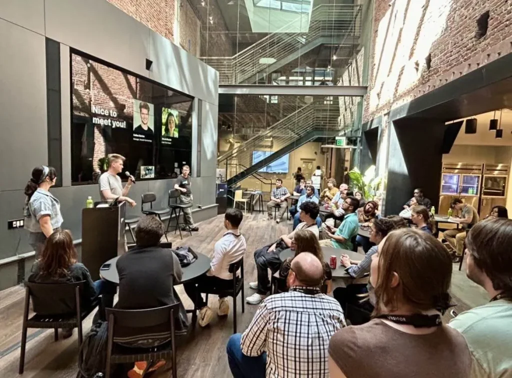 A business presentation in a modern, industrial-style conference room with brick walls and a large screen. A presenter speaks into a microphone, while a diverse audience attentively listens and engages in the session.