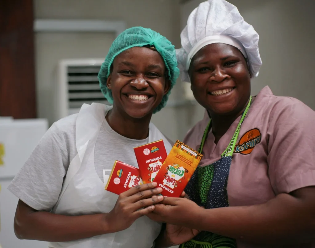 Two smiling women in a chocolate factory, wearing hairnets and chef hats, proudly displaying locally made chocolate bars from Akwaaba brand, representing Ghanaian craftsmanship and entrepreneurship.