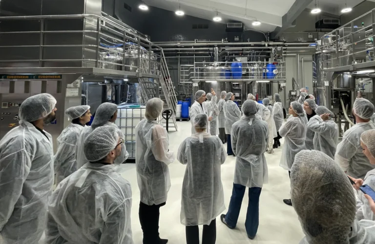 A group of people wearing protective clothing, including hairnets and lab coats, on a guided tour inside a modern industrial facility. The facility features large stainless steel tanks, complex piping systems, and advanced machinery, indicating a production or manufacturing environment.