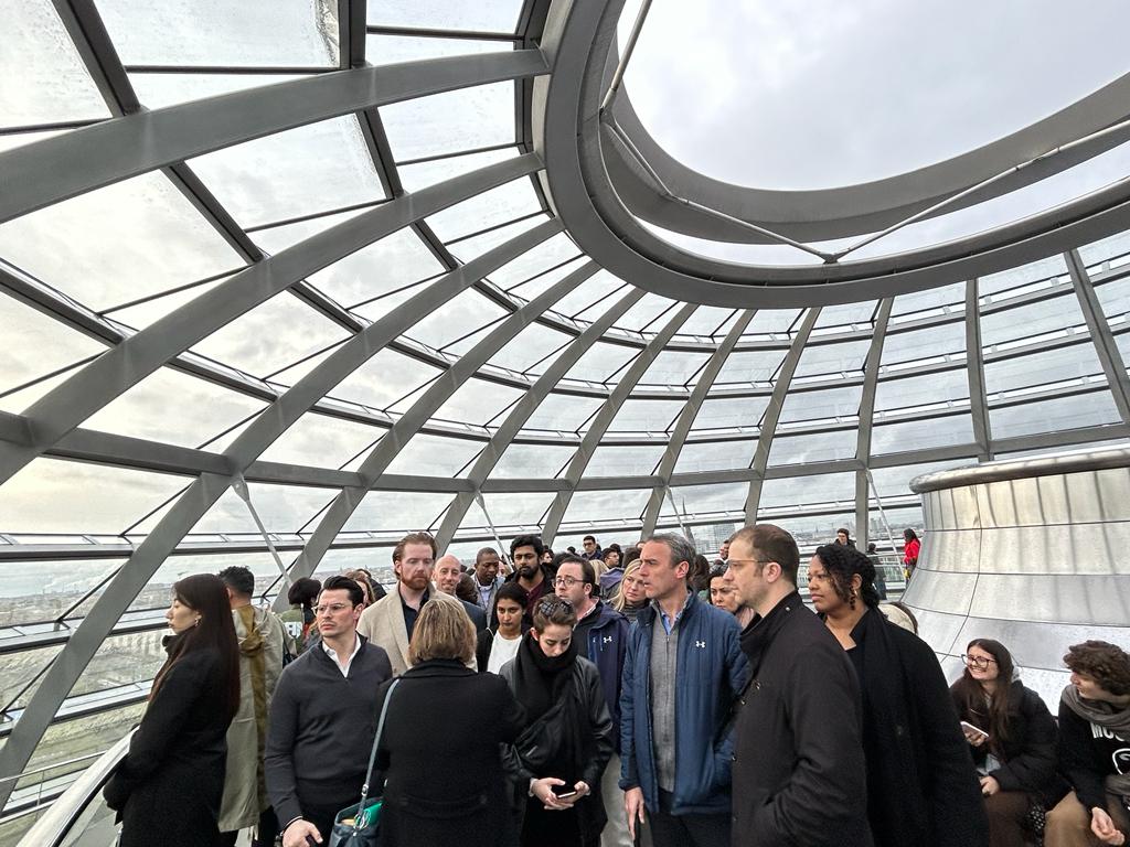 A group of diverse professionals gathered inside the glass dome of the Reichstag Building in Berlin, Germany. The image captures the interior of the dome with its unique spiral architecture, panoramic windows, and a cloudy sky in the background. The group, consisting of men and women of various ages and ethnicities, appears to be on a guided tour or an educational visit. Cornell at Reichstag building.