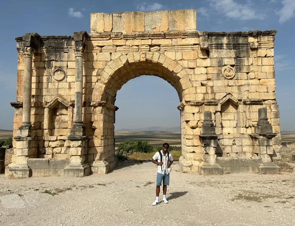 Tourist standing in front of the ancient Arch of Caracalla in Volubilis, Morocco, under a clear blue sky. The well-preserved Roman arch features stone columns and intricate carvings, showcasing the rich historical architecture of the ancient city.