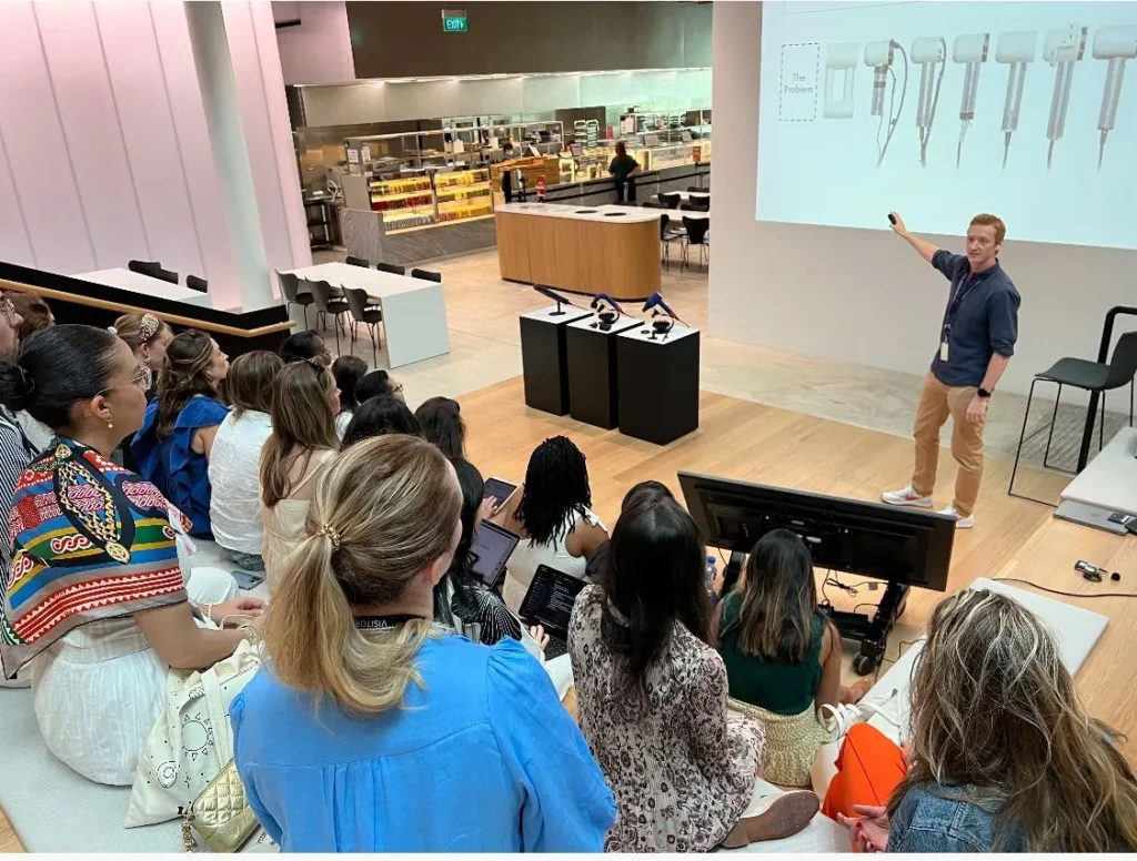 A presenter giving a technical demonstration to a diverse audience in a modern lecture hall, discussing product designs and innovations with visual aids on a large screen.