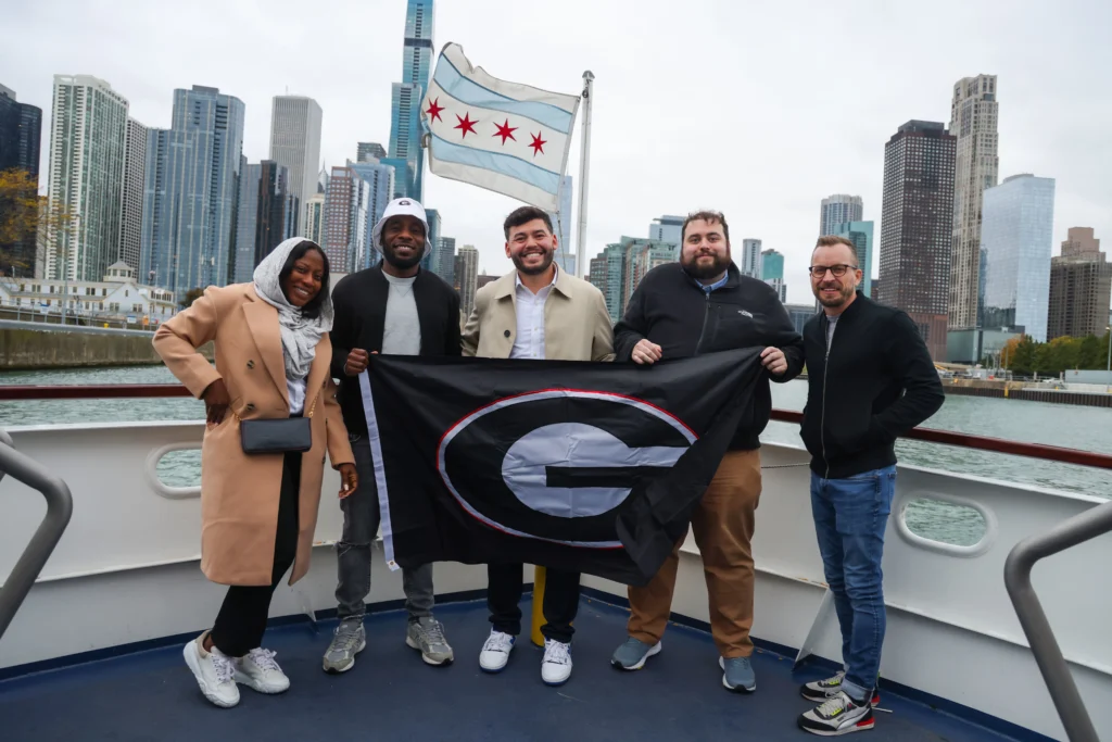 A group of five people posing on a boat in Chicago, holding a University of Georgia flag, with the Chicago skyline and flag visible in the background.