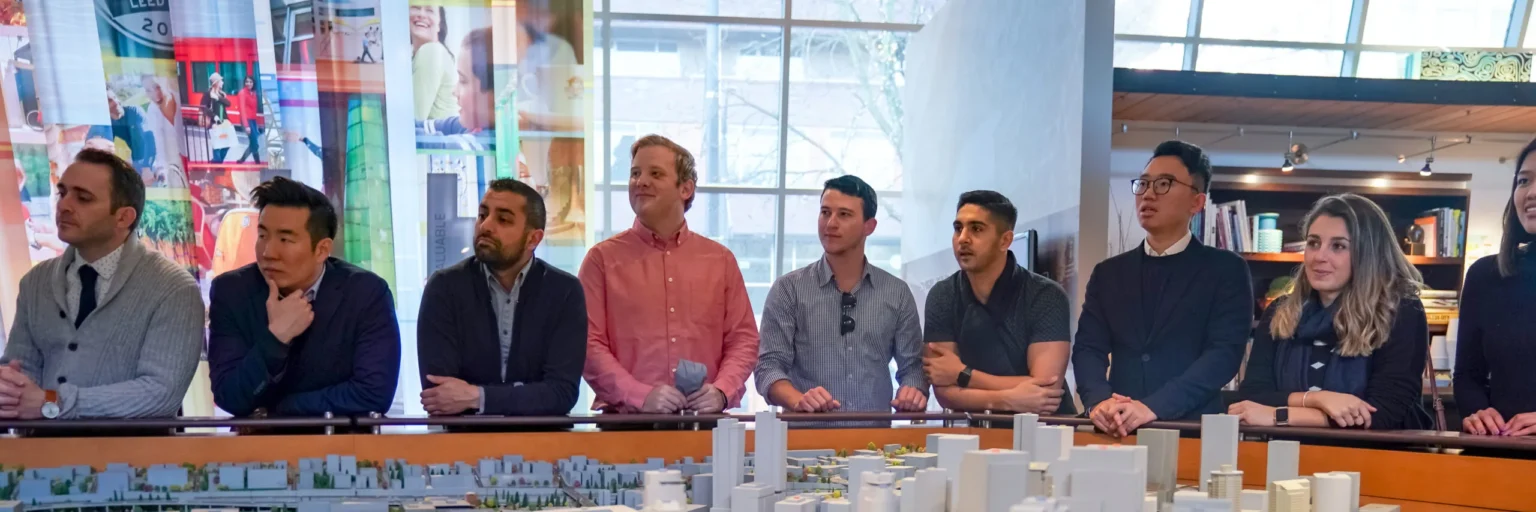 Cornell University. Group of diverse professionals attentively listening during a business presentation in a modern, well-lit conference room, with a detailed architectural model in the foreground.