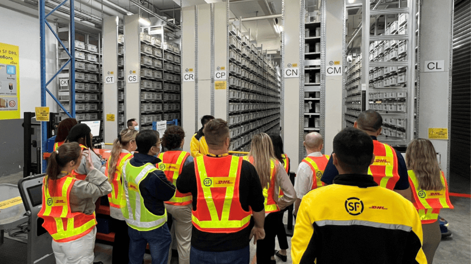 Group of employees wearing high-visibility vests with SF and DHL logos, touring a modern warehouse facility with tall automated storage and retrieval systems filled with inventory bins.