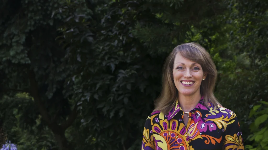 Rebecca Judis. Smiling woman in a colorful patterned blouse standing in front of a lush green background, representing confidence and positivity in an outdoor setting.