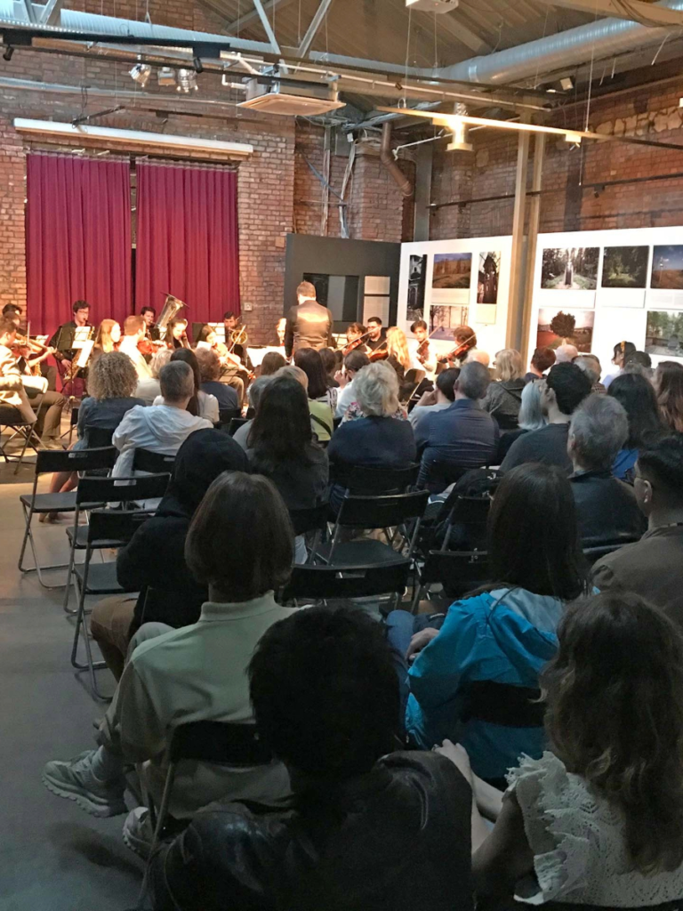 Live orchestra performing in front of an audience in an industrial venue with brick walls and photo exhibits, blending art and music in a cultural event.