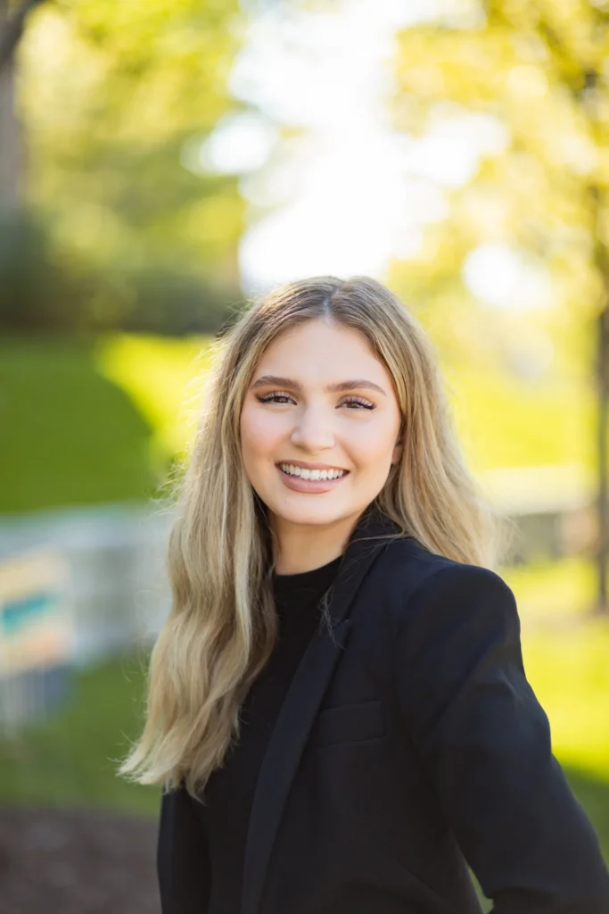 A young woman with long, blonde hair smiles confidently while standing outdoors in a sunlit park. She is dressed in a black blazer and has a friendly, professional demeanor.