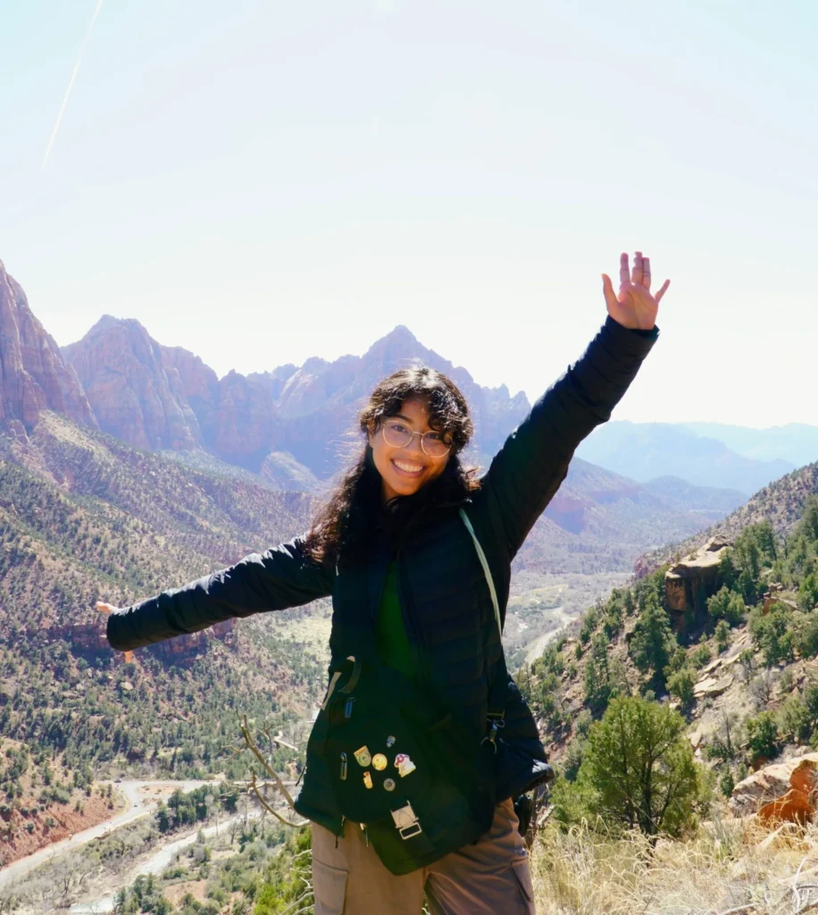 A cheerful hiker stands on a mountain trail, arms outstretched in excitement, with a backdrop of stunning rocky peaks and a river winding through the valley below. She is dressed in hiking gear and wears a crossbody bag adorned with pins.