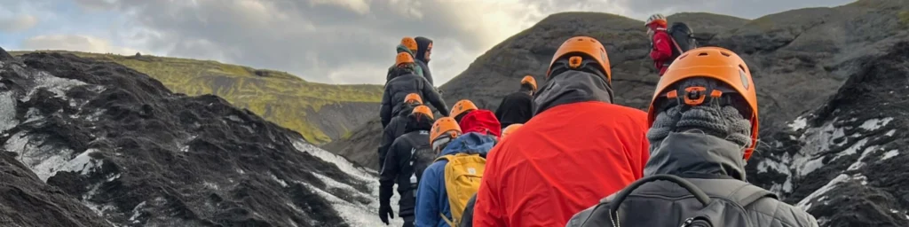 UGA students hiking in Iceland. A group of hikers wearing orange helmets and outdoor gear trekking across a rocky glacier landscape, surrounded by black volcanic rock and patches of ice, under a cloudy sky in Iceland.