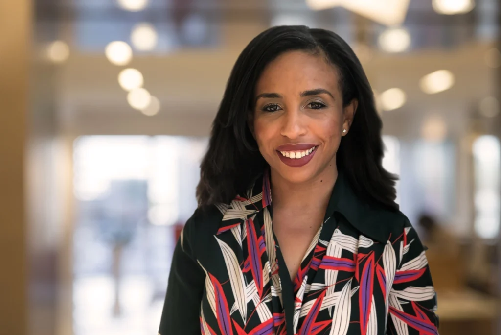 A professional woman smiles warmly in an office setting, wearing a stylish patterned blouse. Her confident expression and polished appearance convey professionalism and approachability.