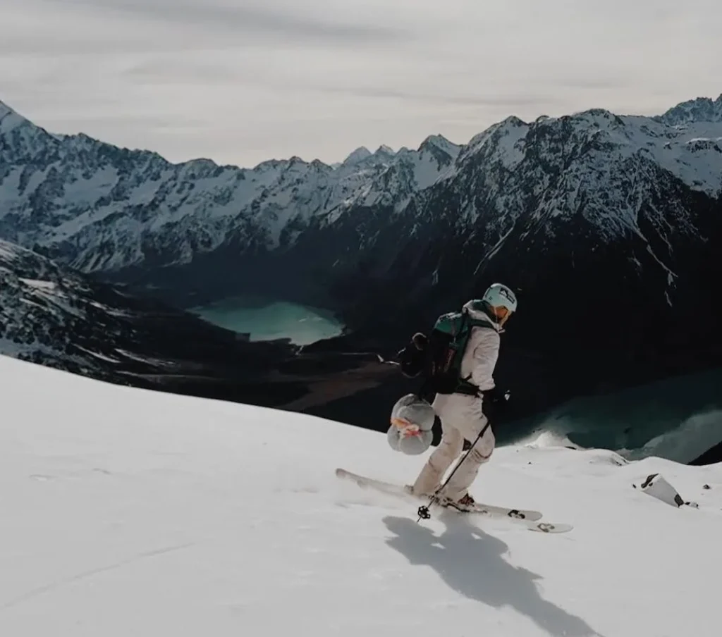 A skier descends a pristine snow-covered mountain slope, surrounded by breathtaking views of rugged snow-capped peaks and serene, glacial lakes below. The skier is equipped with a backpack and gear, embracing the adventure of backcountry skiing.