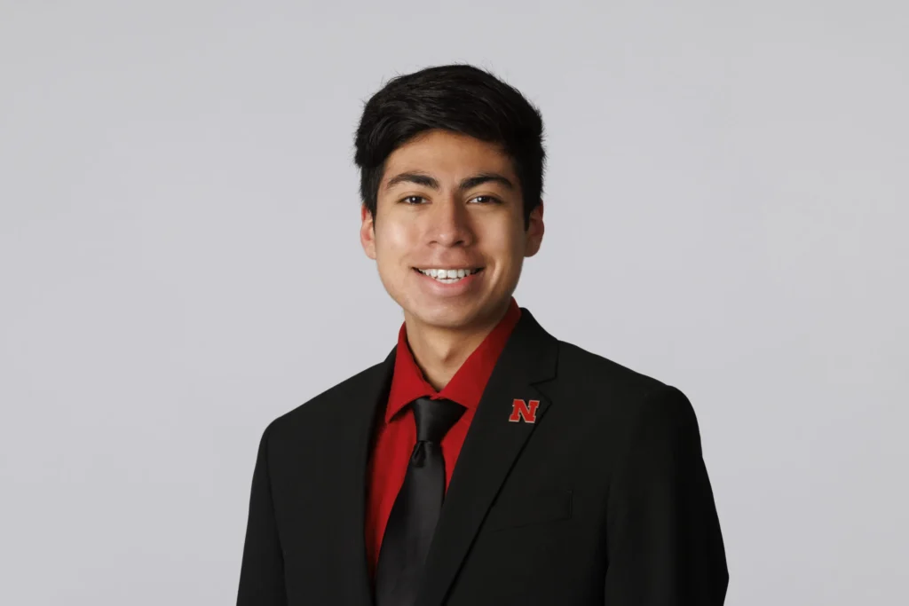 A young professional wearing a black suit and red shirt with a matching red lapel pin featuring the letter "N" smiles confidently in front of a plain gray background.