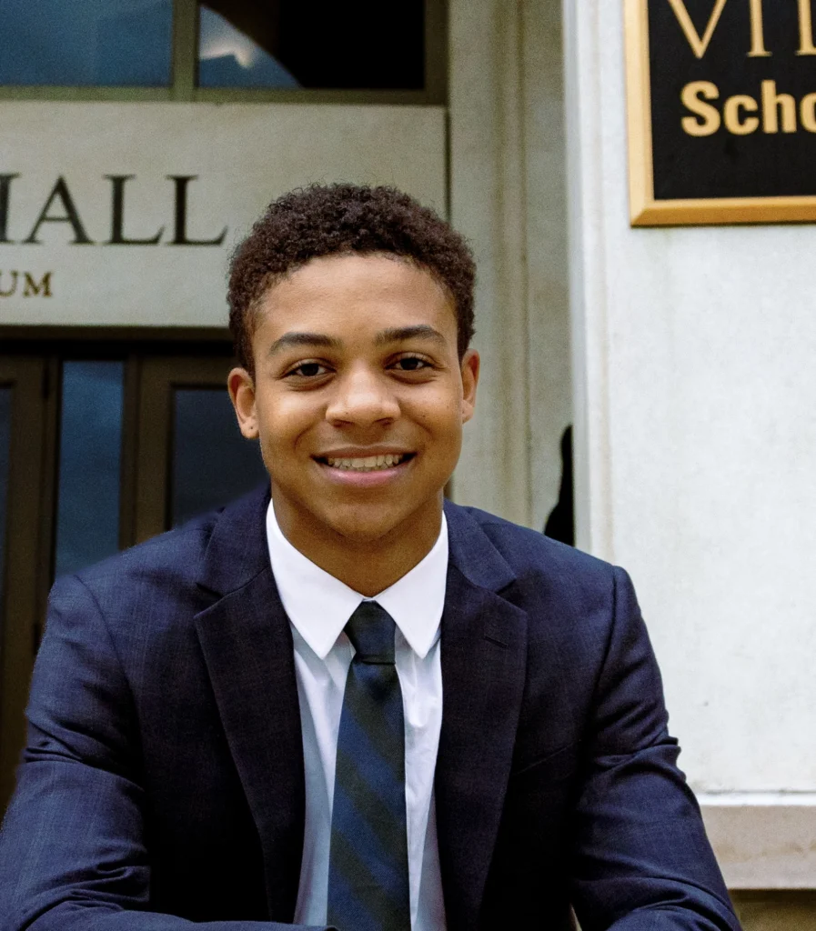 A young professional, dressed in a sharp suit and tie, smiles confidently while sitting outside a building entrance. The backdrop features a sign indicating the building is part of an academic institution.