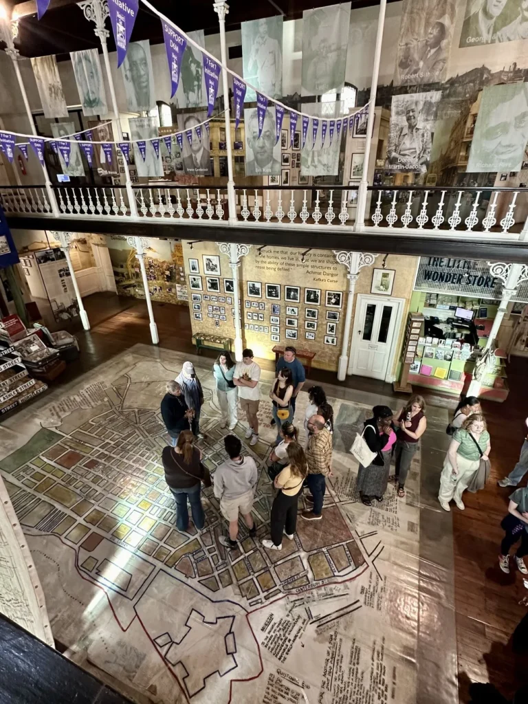 A museum gallery with a group of visitors gathered around a large map of the city, laid out on the floor. The museum walls are covered with historical photographs, flags, and artifacts, offering a rich historical narrative. The balcony above is adorned with banners, and the space exudes an immersive historical ambiance.