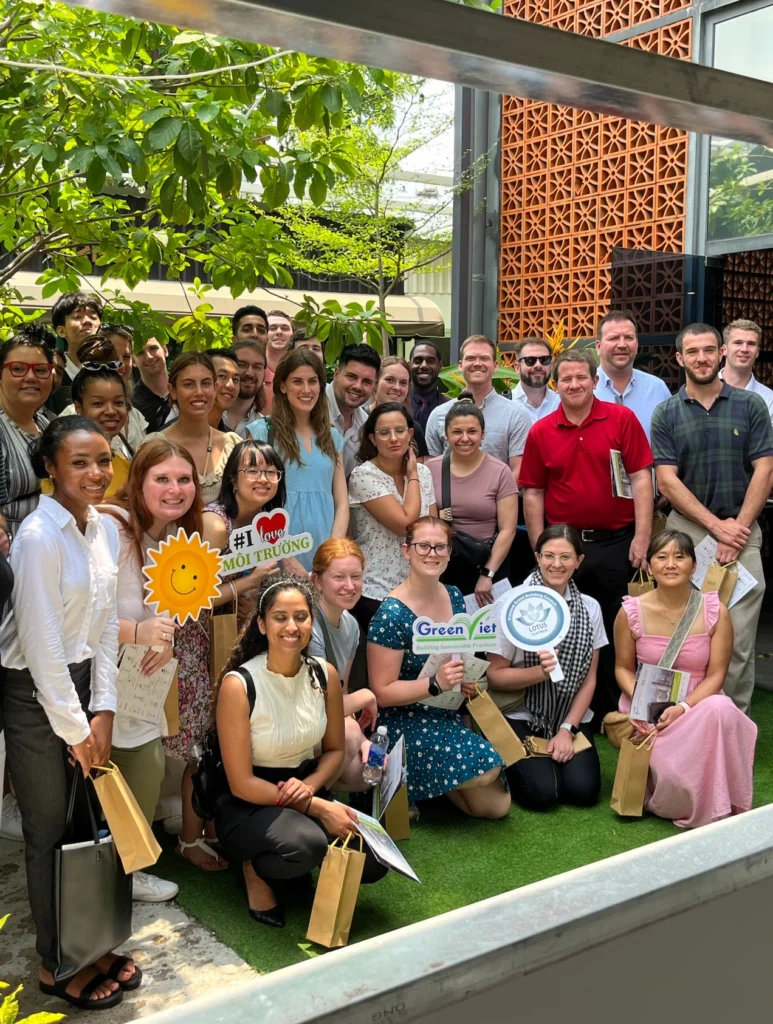 A large group of diverse individuals stands together outside in a garden setting, smiling and holding eco-friendly signs with messages promoting environmental sustainability. Some hold small gift bags, and lush green trees provide a backdrop for the gathering.