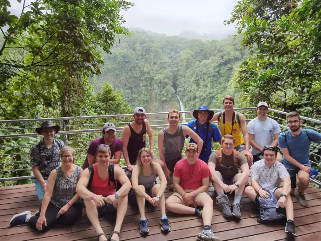 Group of hikers posing in front of a scenic waterfall surrounded by lush greenery.