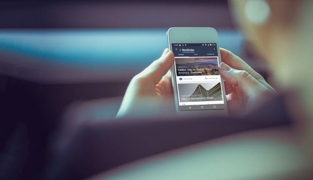 WorldStrides App. Person holding a smartphone with the WorldStrides app open, displaying an itinerary overview for an EMBA trip to Seoul, showcasing educational travel planning and digital itinerary management.